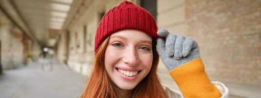 retrato de joven pelirrojo mujer en de punto sombrero y guantes, sonrisas y mira aparte, camina alrededor ciudad en invierno foto