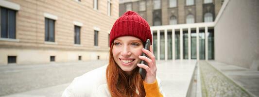 Stylish modern redhead girl talks on mobile phone, makes a telephone call, calling someone on smartphone app from outside, stands on street and smiles photo