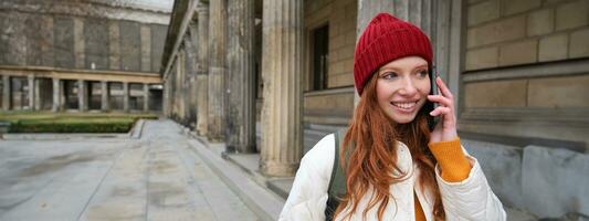 Smiling redhead female tourist talks on mobile phone and walks around city. Happy student in red hat calls friend, stands on street and uses smartphone photo