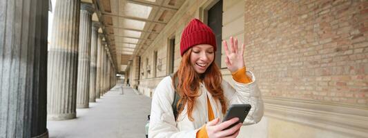 Smiling redhead girl, student says hello, waves hand at smartphone camera, video chats with friend on mobile phone application, walks in city photo