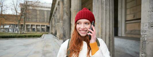 Smiling redhead female tourist talks on mobile phone and walks around city. Happy student in red hat calls friend, stands on street and uses smartphone photo