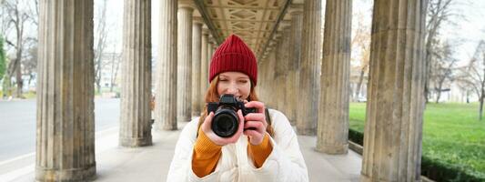 Young redhead female photographer, makes lifestyle shooting in city centre, takes photos and smiles, looks for perfect shot, makes picture