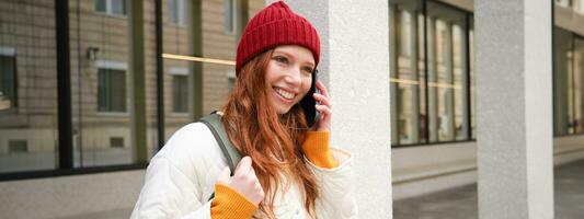 Portrait of smiling ginger girl with red hat, stands on street with backpack, rings someone on phone app, talks on mobile, uses smartphone photo