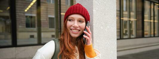 Young people and mobile connection. Happy redhead girl talks on phone, makes telephone call, stands outdoors with backpack and uses smartphone app photo