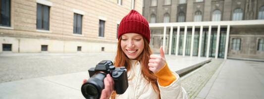 sonriente pelirrojo niña fotógrafo, tomando imágenes en ciudad, hace fotos al aire libre en profesional cámara