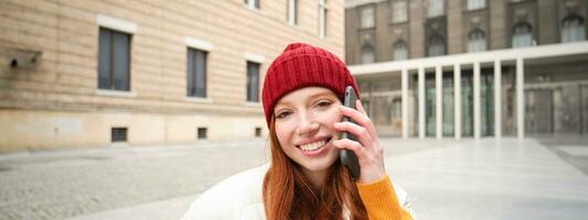 móvil conexión y personas concepto. contento pelirrojo mujer en sombrero, negociaciones en móvil teléfono, haciendo teléfono llamar, utilizando aplicación a llamada extranjero foto