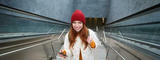 Portrait redhead girl tourist, goes up stairs with smartphone, follows route on mobile phone app, holds backpack and smiles photo