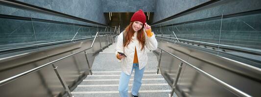 Portrait redhead girl tourist, goes up stairs with smartphone, follows route on mobile phone app, holds backpack and smiles photo