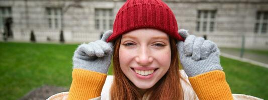 Close up portrait of beautiful redhead woman in red knitted hat, warm gloves, smiling and looking happy at camera, sitting in park photo