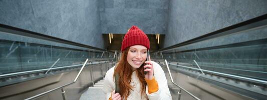 retrato de contento pelirrojo mujer caminando alrededor pueblo con teléfono inteligente, vocación alguien, hablando en móvil teléfono al aire libre foto