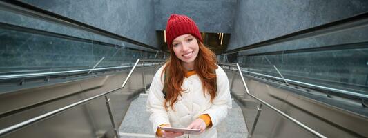 Beautiful redhead female model posing in city, walking up stairs with digital tablet, using gadget to plan her route, reading while going somewere photo