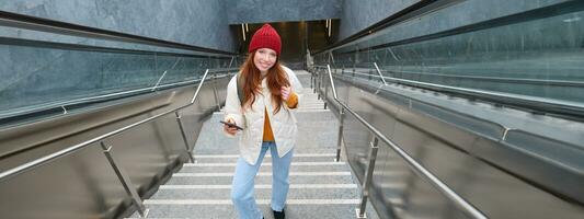 Portrait redhead girl tourist, goes up stairs with smartphone, follows route on mobile phone app, holds backpack and smiles photo