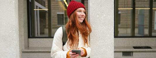 Beautiful redhead girl with smartphone, standing on street, holding mobile phone, using taxi app, connects to public wifi and uses internet, sends a text message photo