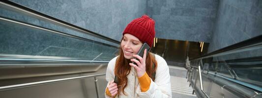 Portrait of young redhead woman walks around city, goes up stairs with mobile phone, talks on smartphone and smiles photo