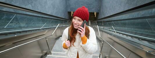 retrato de joven pelirrojo mujer camina alrededor ciudad, va arriba escalera con móvil teléfono, negociaciones en teléfono inteligente y sonrisas foto