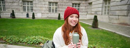 Smiling redhead girl rests in park, sits on bench with backpack, drinks from thermos, enjoys hot drink from flask and looks relaxed photo