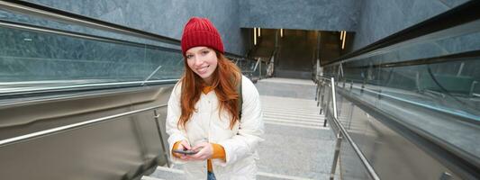 Smiling girl walking on stairs, using mobile phone, texting message on her way, using smartphone app map, follows route to destination place photo