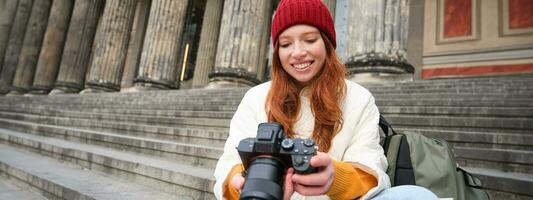 Portrait of young photographer girl, sits on stairs with professional camera, takes photos outdoors, making lifestyle shooting