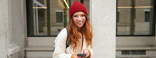 Stylish urban girl using mobile phone app, standing in city, waiting for taxi, looking at smartphone application, texting message photo