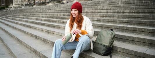 Smiling tourist, girl sits on stairs, rests on staircase, takes thermos from backpack, drinks hot coffee from flask photo