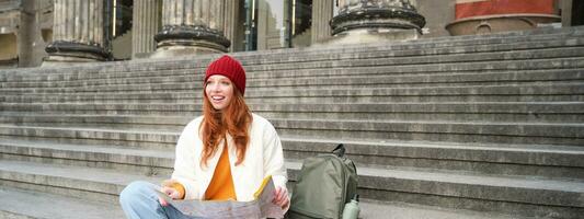 Portrait of young redhead woman, tourist sits with paper map and looks for a route to tourism attraction, rests on stairs outdoors photo