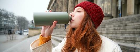 Young redhead female tourist rests during her trip, opens thermos and drinks hot tea, having a break after sightseeing photo