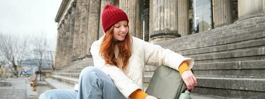 Smiling tourist, girl sits on stairs, rests on staircase, takes thermos from backpack, drinks hot coffee from flask photo