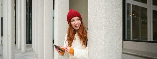 Stylish urban girl using mobile phone app, standing in city, waiting for taxi, looking at smartphone application, texting message photo