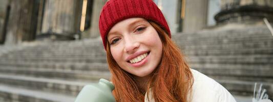 Young redhead female tourist rests during her trip, opens thermos and drinks hot tea, having a break after sightseeing photo