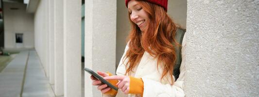 Stylish urban girl using mobile phone app, standing in city, waiting for taxi, looking at smartphone application, texting message photo