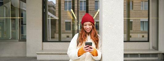 Stylish urban girl using mobile phone app, standing in city, waiting for taxi, looking at smartphone application, texting message photo