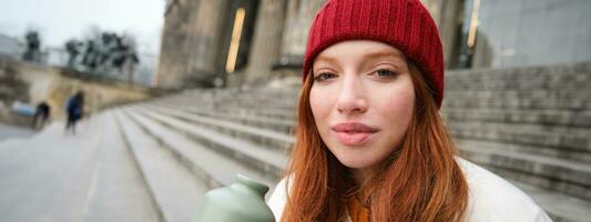 Young redhead female tourist rests during her trip, opens thermos and drinks hot tea, having a break after sightseeing photo