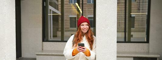Beautiful redhead girl with smartphone, standing on street, holding mobile phone, using taxi app, connects to public wifi and uses internet, sends a text message photo