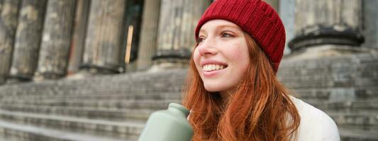 Smiling redhead girl in red hat, sits on stairs near building with flask, drinks water and looks happy photo