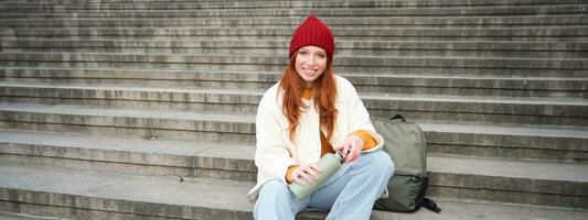 Smiling tourist, girl sits on stairs, rests on staircase, takes thermos from backpack, drinks hot coffee from flask photo