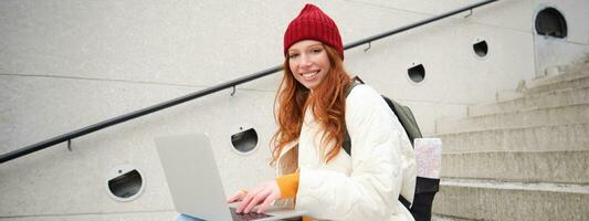 Portrait of young girl traveller, sitting with backpack and map of city, working on laptop, connecting to public wifi and sitting on stairs outdoors, using internet to book hotel room photo