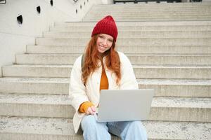 Smiling redhead girl, student sits on stairs outdoors and uses laptop, connects to public wifi in city and works on project, uses internet on computer photo