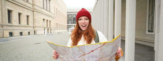 hermosa pelirrojo mujer, turista con ciudad mapa, explora Turismo histórico punto de referencia, caminando alrededor antiguo ciudad, sonriente felizmente foto