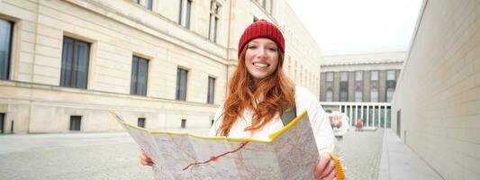 hermosa pelirrojo mujer, turista con ciudad mapa, explora Turismo histórico punto de referencia, caminando alrededor antiguo ciudad, sonriente felizmente foto