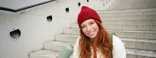Happy redhead woman, tourist with thermos, drinks her hot tea, coffee from travel flask, restests during her travelling in city and sightseeing photo