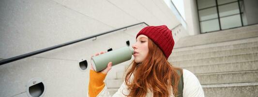 Smiling traveler, redhead girl tourist sits on stairs with flask, drinks hot coffee from thermos while travelling and sightseeing around foreign city, sits on stairs and rests photo