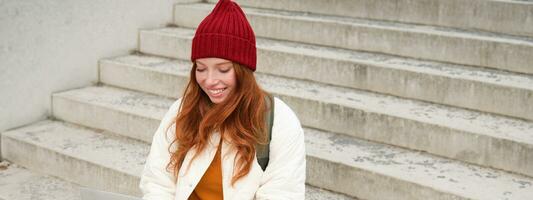 Smiling redhead girl, young woman typing on laptop keyboard, sitting outdoors on stairs with computer, working remote, doing her homework on fresh air photo