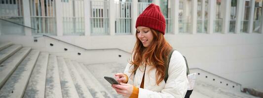 Smiling redhead girl, student tourist walks around city, goes up the stairs, looks at mobile phone map to get around town, sends message on smartphone photo