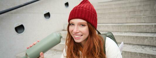 Happy redhead woman, tourist with thermos, drinks her hot tea, coffee from travel flask, restests during her travelling in city and sightseeing photo