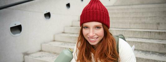 Smiling traveler, redhead girl tourist sits on stairs with flask, drinks hot coffee from thermos while travelling and sightseeing around foreign city, sits on stairs and rests photo