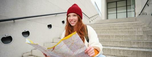 joven sonriente pelirrojo chica, turista se sienta en escalera al aire libre con ciudad papel mapa, mirando para dirección, viajero mochilero explora ciudad y mira para Turismo foto