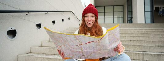 hermosa niña turista se sienta en escalera con ciudad mapa, planes su viaje, mira para dirección mientras de viaje alrededor ciudad, búsquedas ruta para Turismo foto