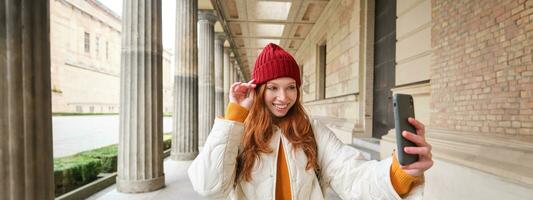 Stylish redhead girl tourist, takes selfie in front of tourism attraction, makes photo with smartphone, looks at mobile camera and poses