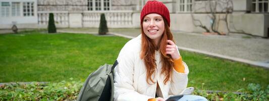 Redhead girl, female student sits with mobile phone on bench in parj, leans on her backpack. Woman browsing social media app feed on her smartphone photo