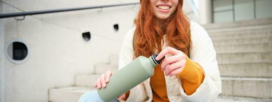 Smiling traveler, redhead girl tourist sits on stairs with flask, drinks hot coffee from thermos while travelling and sightseeing around foreign city, sits on stairs and rests photo